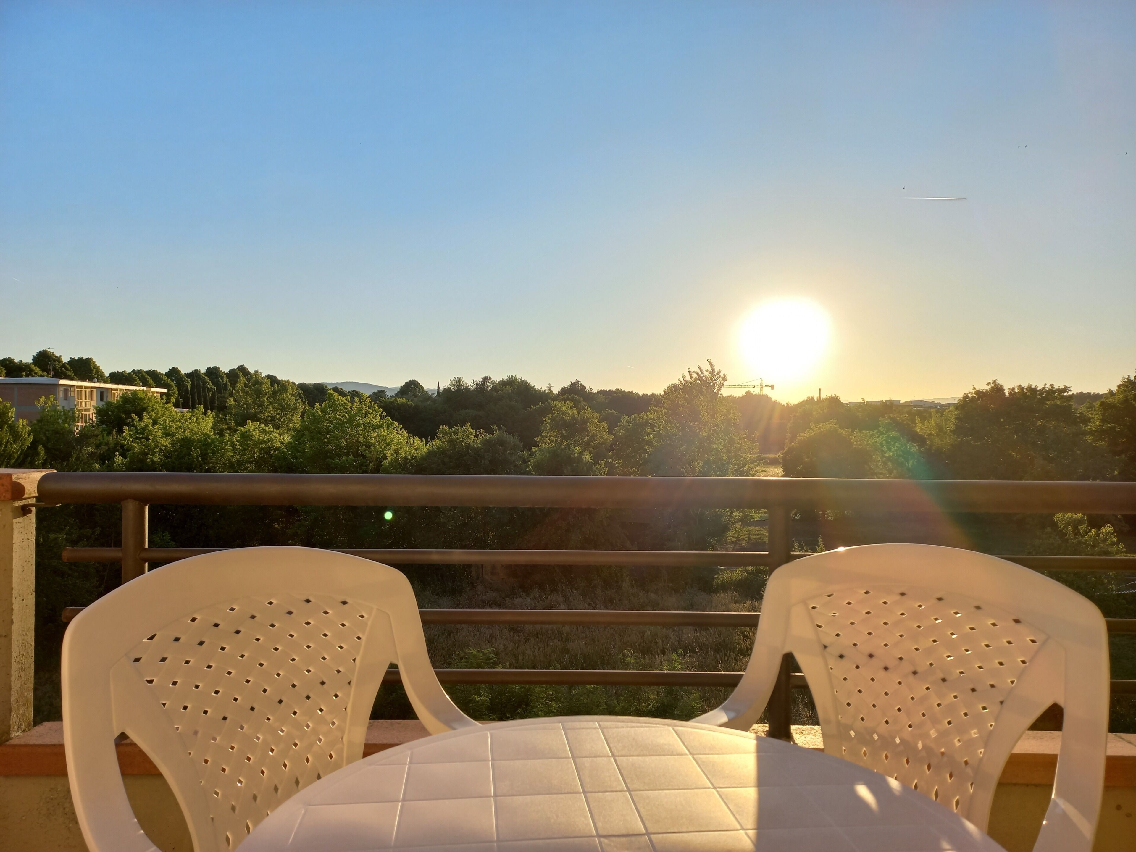A balcony with two white plastic chairs and a table, overlooking a landscape with trees and a building in the distance.. Appartamento vista parco