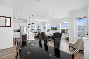 Una cocina moderna con armarios blancos, un comedor con mesa y sillas negras, y una sala de estar con sofá y televisor.