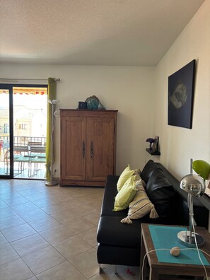 A living room with a black leather sofa, a wooden cabinet, a floor lamp, and a dining area with chairs and a table. - Apartment Calvi center Terrace air-conditioned (Calvi)