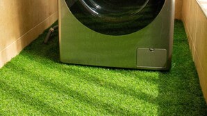 Une machine à laver à chargement frontal se trouve dans une buanderie, dont le sol est recouvert d’un gazon artificiel vert. On y voit également un robinet et une étagère contenant des produits de nettoyage.