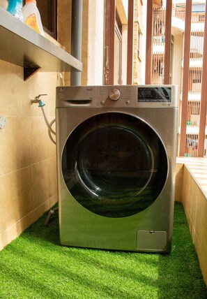 Une machine à laver à chargement frontal se trouve dans une buanderie, dont le sol est recouvert d’un gazon artificiel vert. On y voit également un robinet et une étagère contenant des produits de nettoyage.