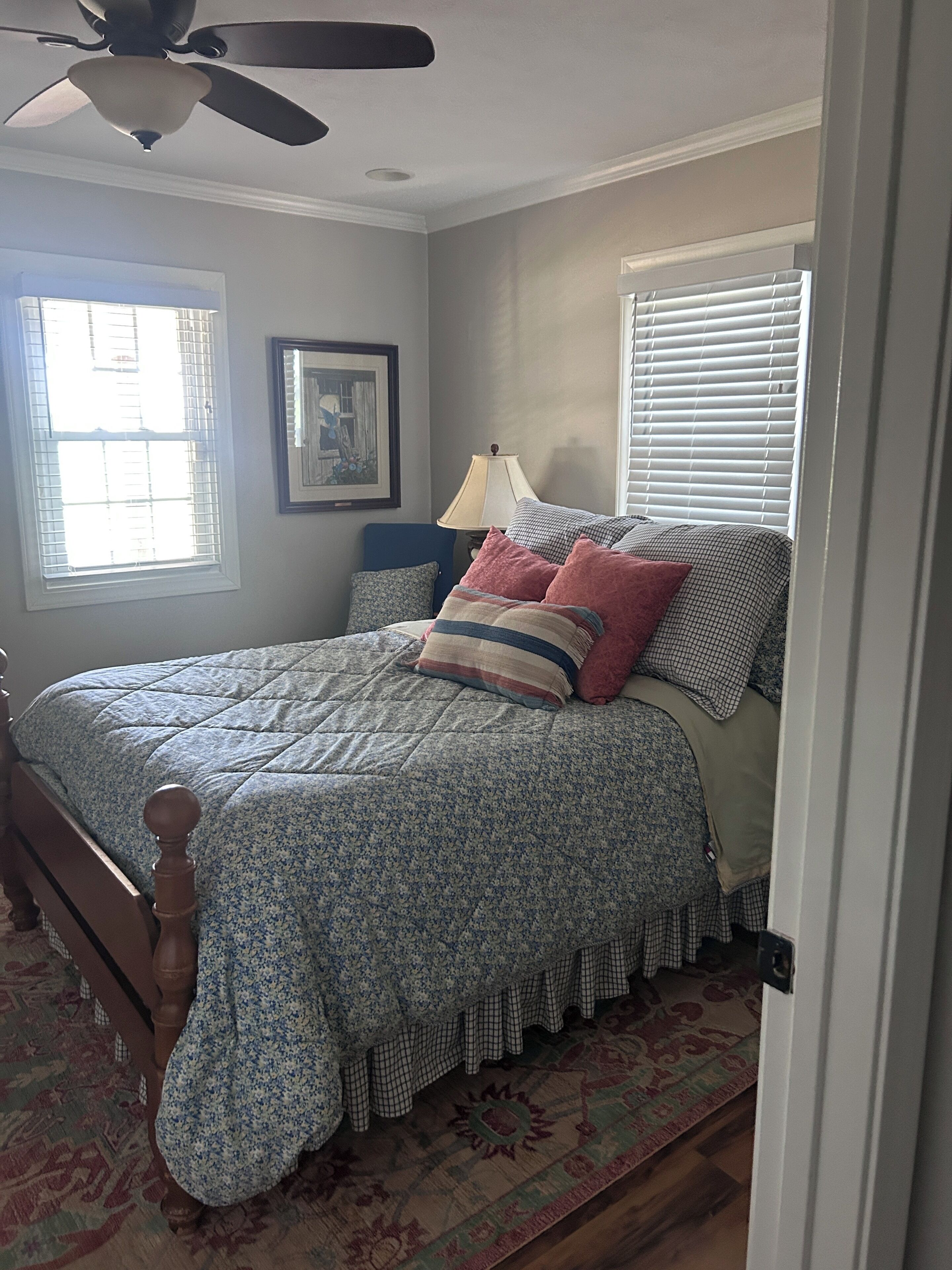 A bedroom with a bed, a ceiling fan, a window with blinds, a lamp, and a framed picture on the wall.