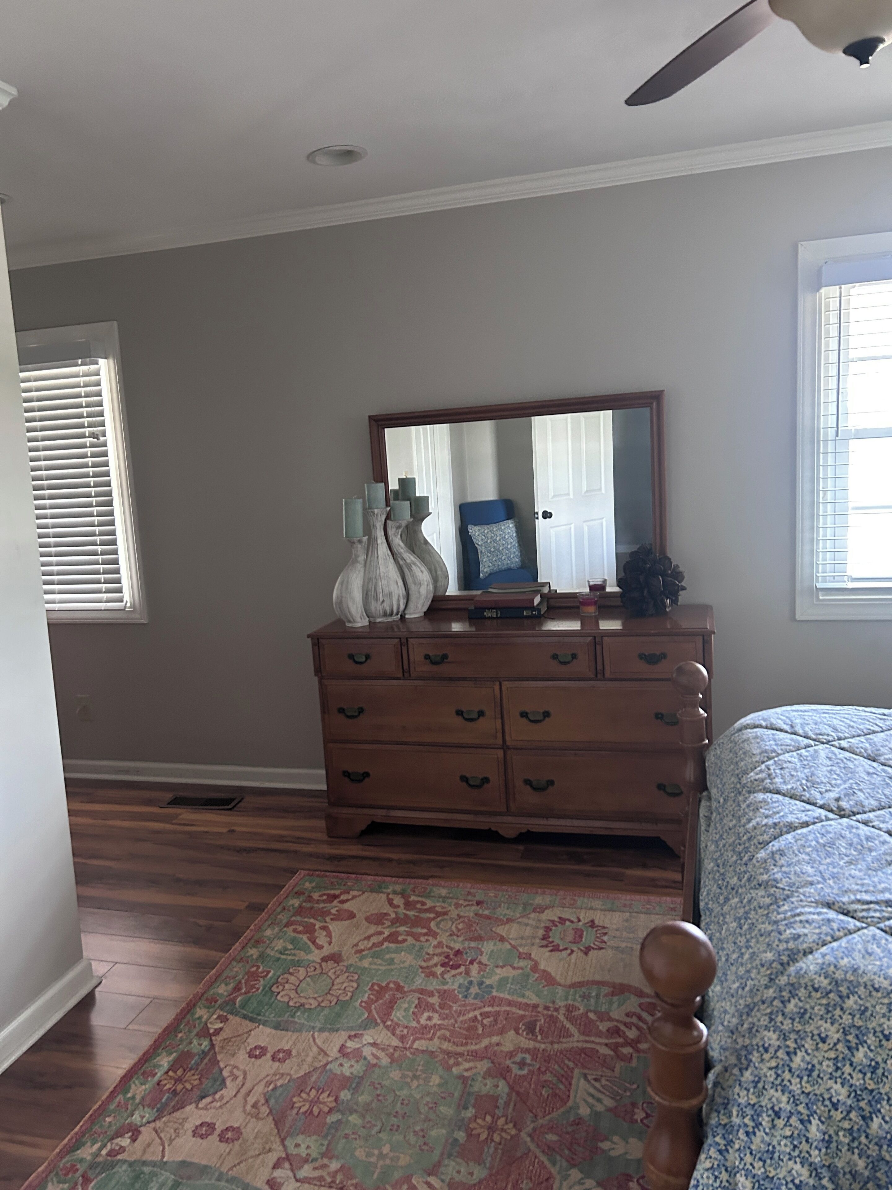 Une chambre avec une commode en bois, un grand miroir, un tapis à motifs et un lit recouvert d’une couvre-lit à motifs bleus et blancs.