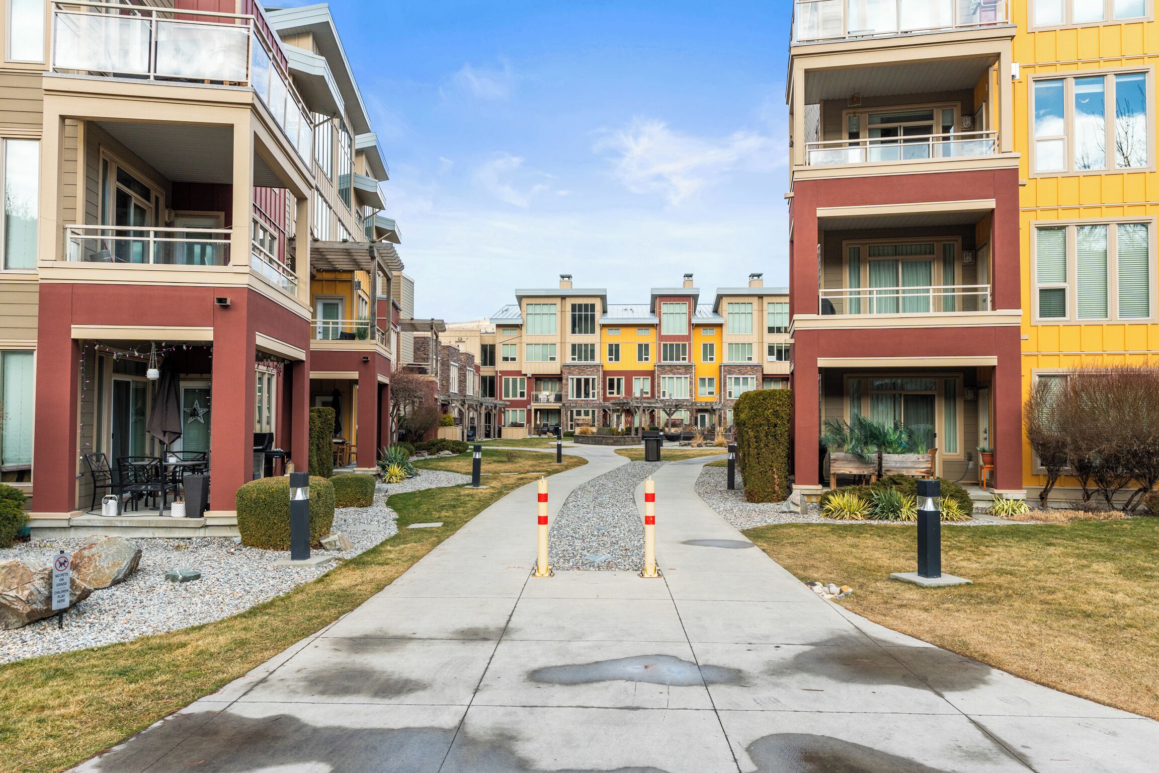 A row of multi-story residential buildings with balconies, a central pathway, and small landscaping.