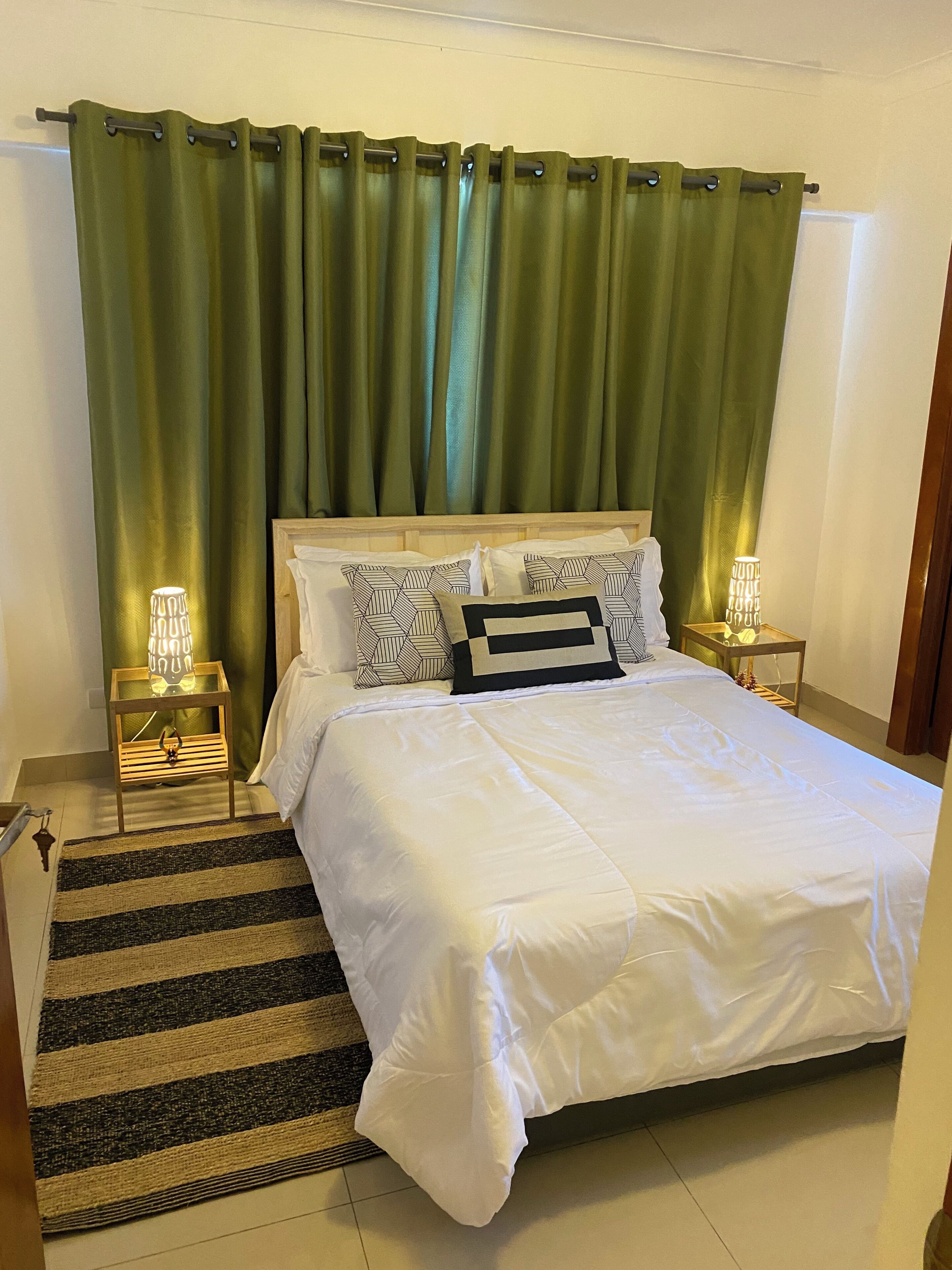 Una cama bien hecha con una colcha blanca y almohadas estampadas, una cortina verde y dos lámparas de noche sobre mesitas de madera.