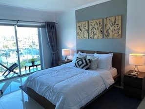 Ein Hotelzimmer mit einem Bett, zwei Nachttischlampen, einem Fenster mit Vorhängen, einem Balkon und Kunstwerken an der Wand.