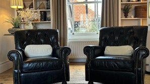 Ein Zimmer mit zwei schwarzen Ledersesseln, einem Schachbrett, einem fenster mit Vorhängen und einem Bücherregal mit verschiedenen Gegenständen.