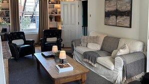 Ein Wohnzimmer mit einer Couch, zwei Sesseln, einem Couchtisch und einer Kandelaber. Ein Bücherregal und eine Tür zu einem anderen Raum sind ebenfalls vorhanden.