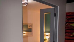 A narrow hallway leading to a bedroom with a bed, a bedside table, and a wall-mounted lamp. - Ocean View! Jacuzzi! Outdoor Living! Centrally Located! (Arecibo)