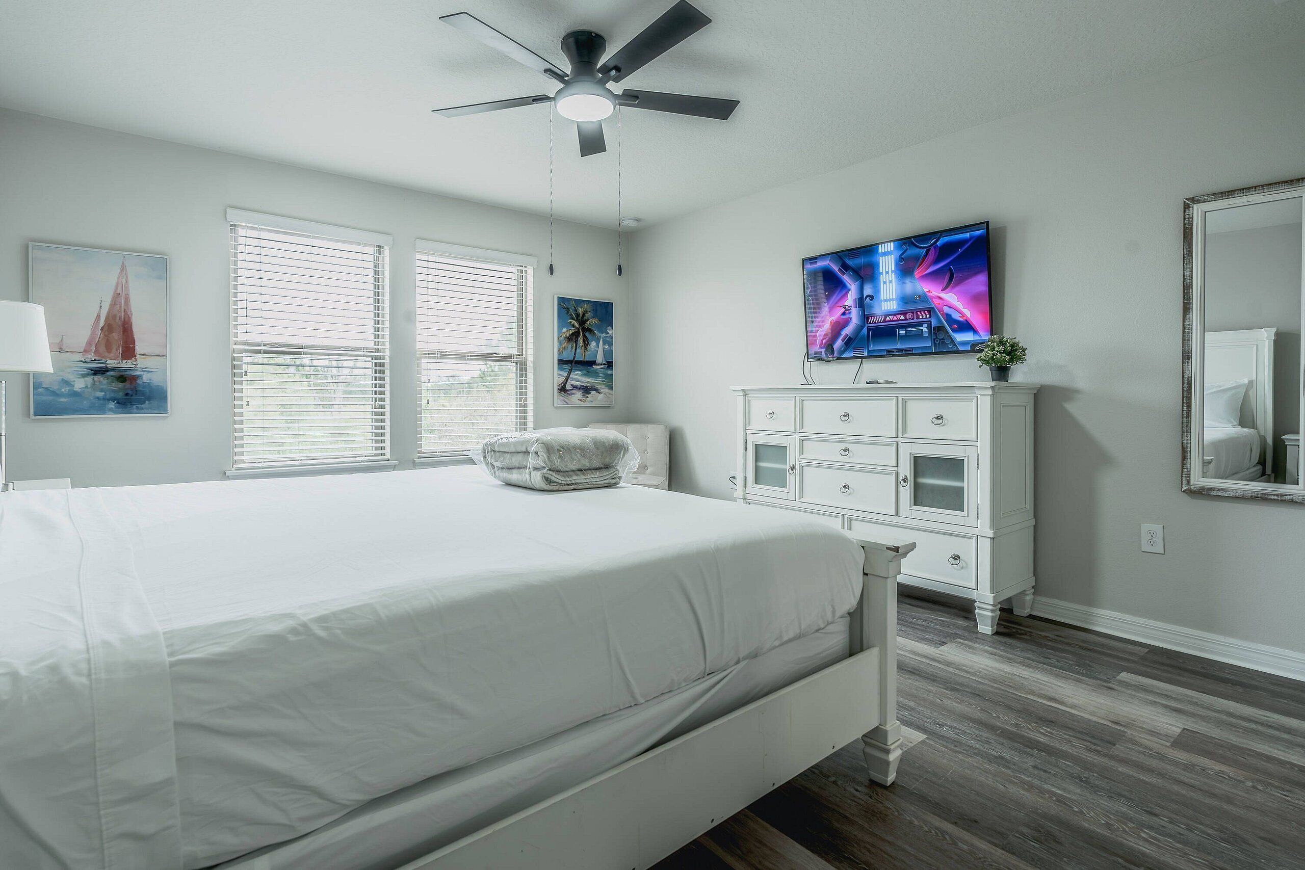 A bedroom with a bed, a ceiling fan, a TV, a dresser, and a window with blinds.