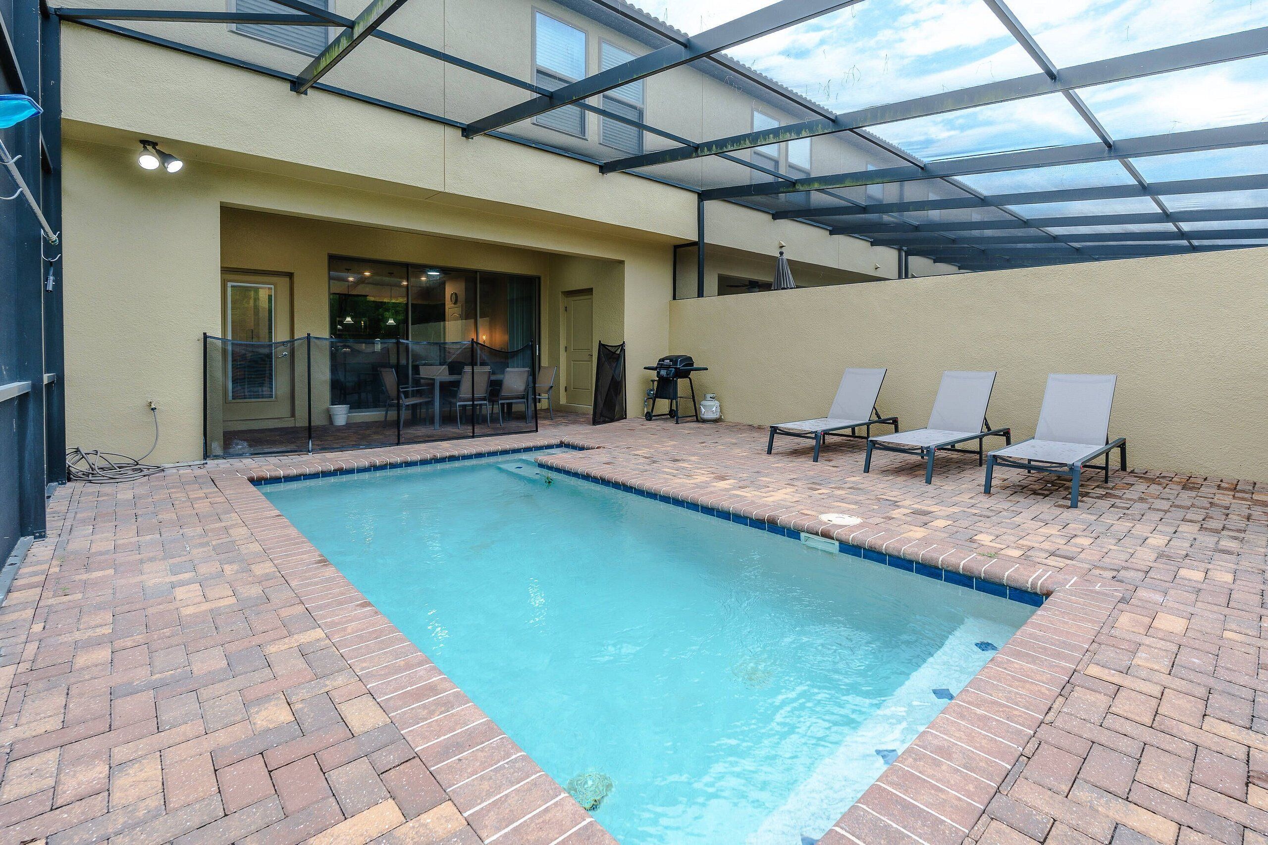 A swimming pool with a brick border, a covered patio area with chairs, and a glass-enclosed room with a door.