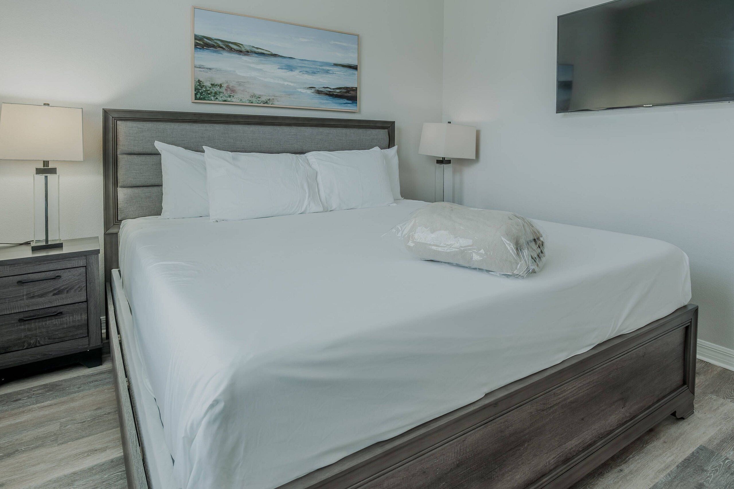 A neatly made bed with white linens, a wooden headboard, a bedside table with a lamp, and a painting above the bed.