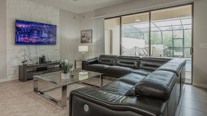 A modern living room with a sectional sofa, a glass coffee table, a flat-screen TV, and a ceiling fan. - 5BR Solterra Home Near Disney With Pool (Davenport)