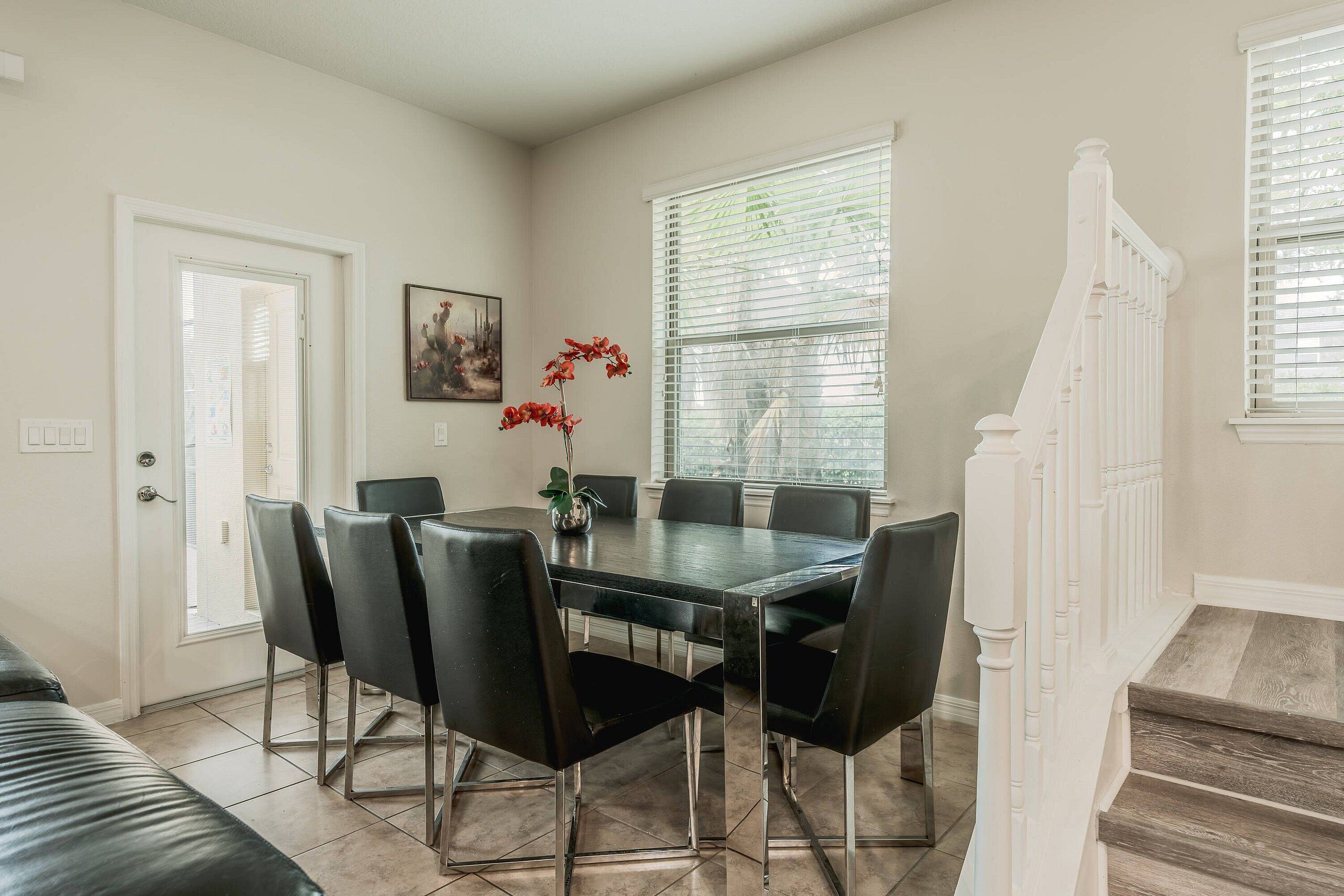 A dining area with a table and chairs, a vase with flowers, a painting on the wall, and a staircase.