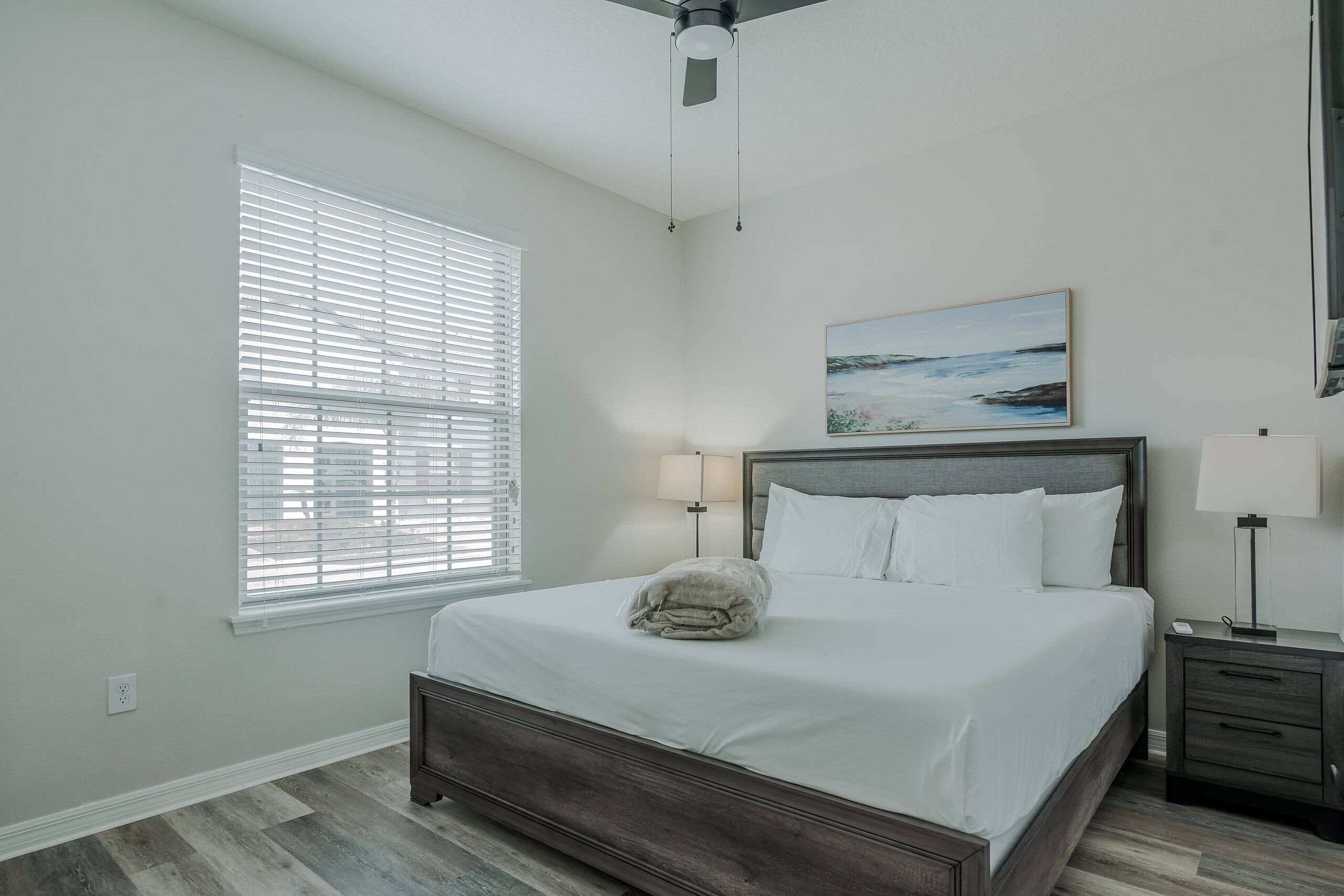 A bedroom with a bed, a window with blinds, a ceiling fan, a bedside table with a lamp, and a painting on the wall.