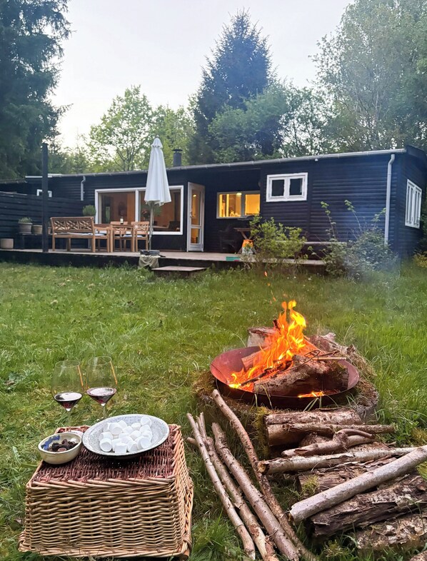 A cozy outdoor setting with a fire pit, wicker table with dishes, and a small house with a deck and outdoor seating. - Cozy holiday home near Tisvilde Hegn (Tibirke Sand, Frederiksværk)