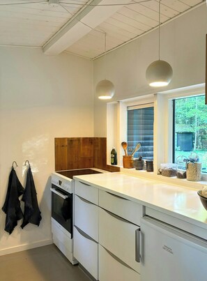 A modern kitchen with white cabinetry, a built-in oven, a window with a view of greenery, and hanging kitchen towels. - Cozy holiday home near Tisvilde Hegn (Tibirke Sand, Frederiksværk)