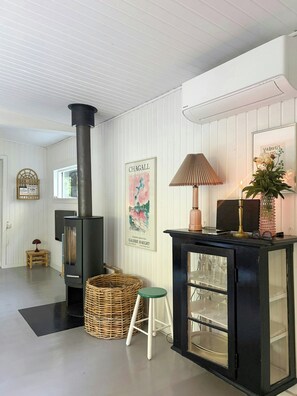 A room with a wood-burning stove, a wicker basket, a small stool, a glass cabinet with wine glasses, and a framed artwork on the wall. - Cozy holiday home near Tisvilde Hegn (Tibirke Sand, Frederiksværk)