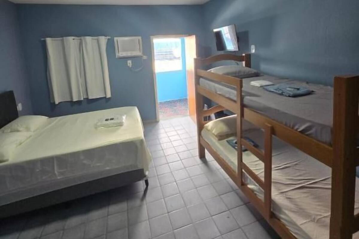 Habitación de residencia estudiantil con dos literas, un televisor en la litera superior, una ventana con cortinas y una puerta que da al exterior.