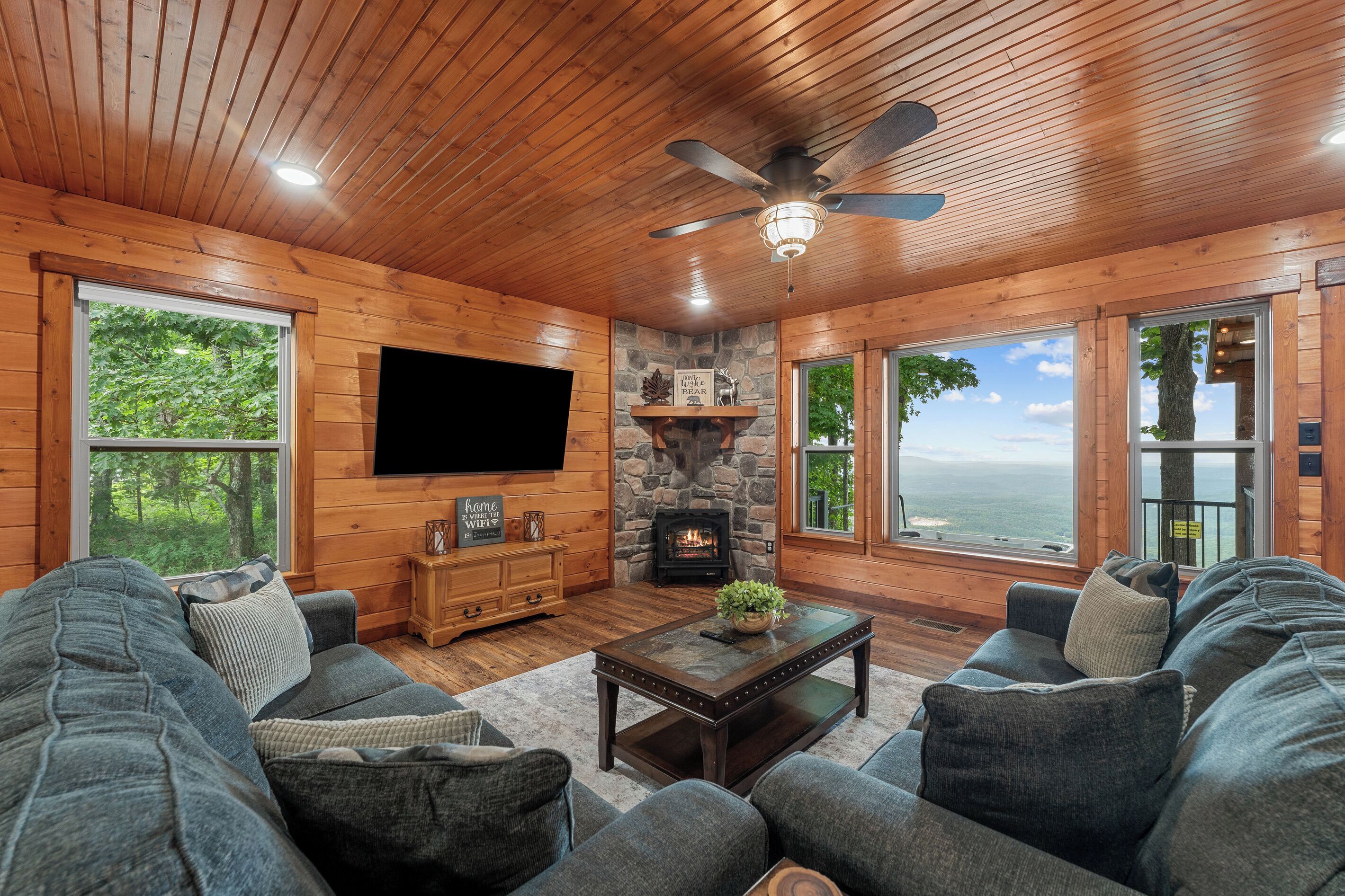 Una sala de estar acogedora con chimenea de piedra, paredes de madera, ventilador de techo y ventanas amplias con vistas panorámicas.