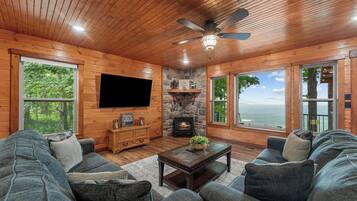 Una sala de estar acogedora con chimenea de piedra, paredes de madera, ventilador de techo y ventanas amplias con vistas panorámicas.