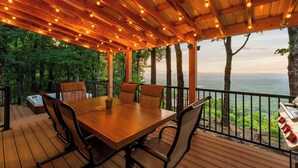An outdoor wooden deck with a table and chairs, surrounded by trees and a view of the landscape.