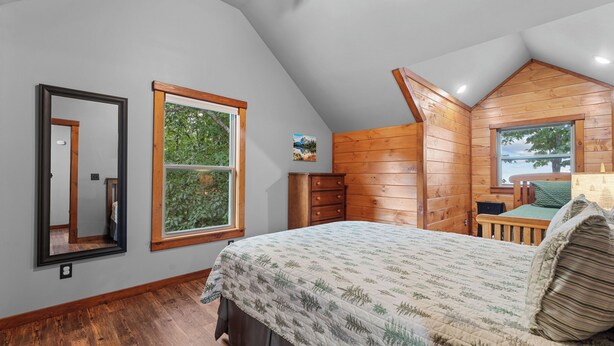 A bedroom with a sloped ceiling, wooden walls, a bed with a patterned bedspread, a dresser, and a window with a view of greenery.