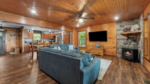 A cozy cabin interior with a wooden ceiling, a stone fireplace, a large sofa, and a dining area.