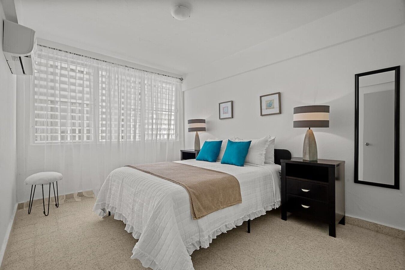 A bedroom with a bed, two bedside tables, a chair, a window with blinds, and a door.