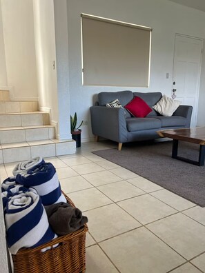 A living room with a grey sofa, a coffee table, a staircase, and a basket of folded towels. - Tropical Tamarindo Condo; 5-Min Walk to Beach (Tamarindo)