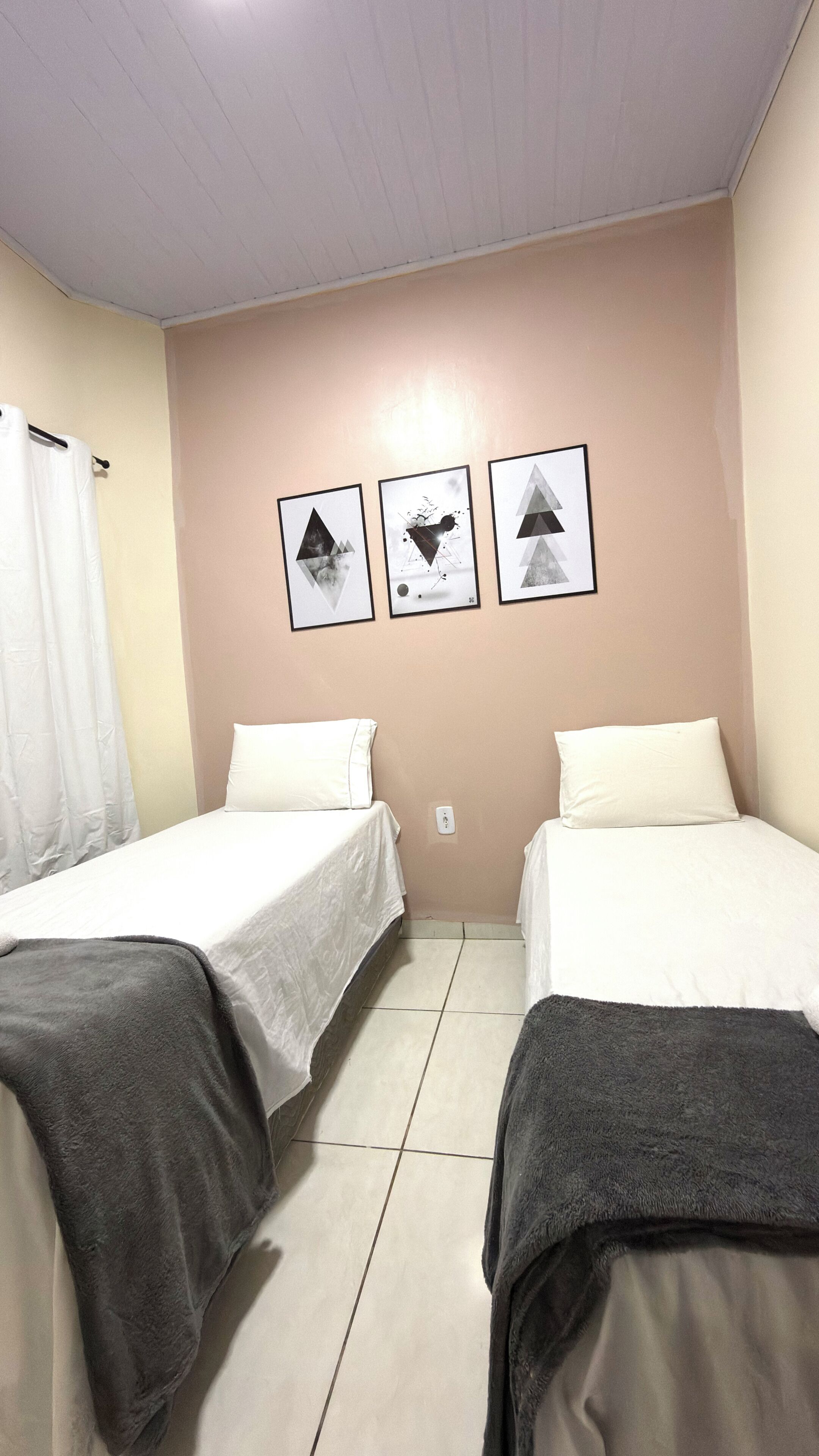 Quarto com duas camas, uma cortina branca e três quadros na parede.