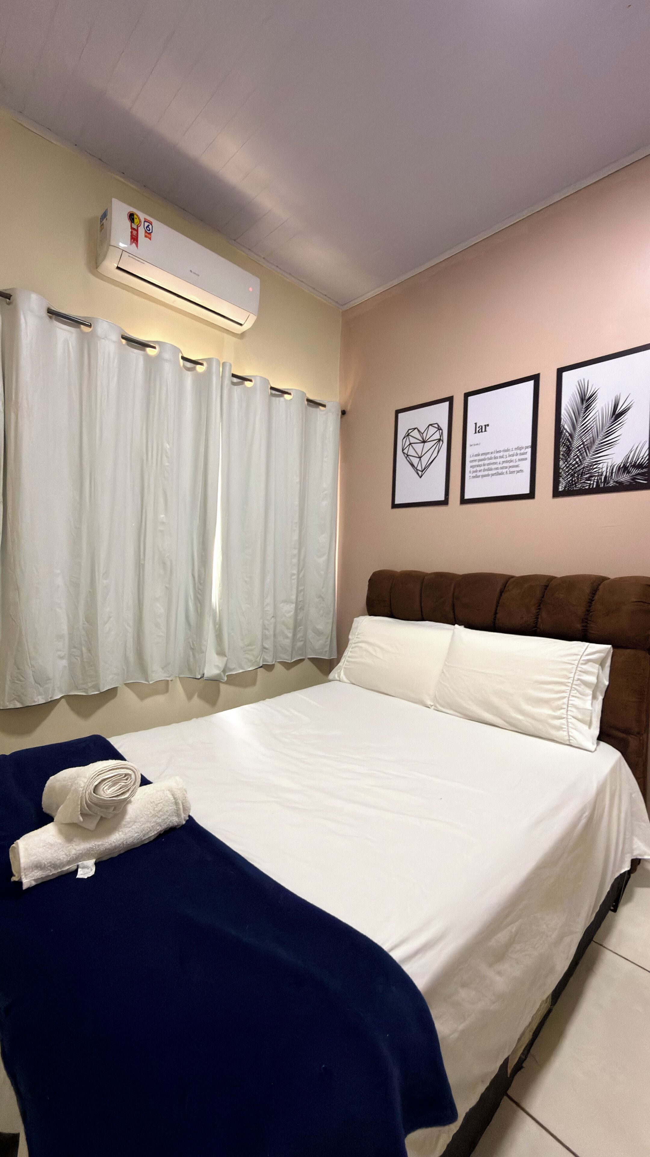 A hotel room with a bed, white curtains, a wall-mounted air conditioner, and framed artwork.