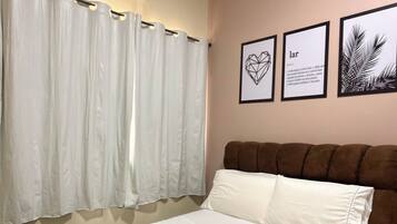 A hotel room with a bed, white curtains, a wall-mounted air conditioner, and framed artwork.