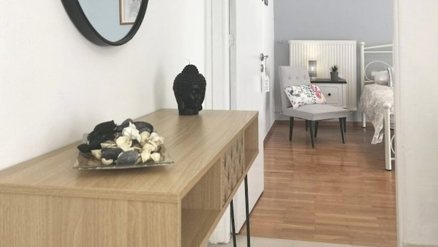 Ein Zimmer mit einem Holztisch, einem schwarzen Spiegel und einer Buddha-Statue. Es gibt ein Bett mit einem weißen Kopfteil sowie einen kleinen Tisch mit einem Stuhl.