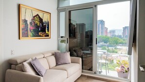 Ein modernes Wohnzimmer mit einem beigefarbenen Sofa, einer Glastür, die auf einen Balkon führt, und Blick auf die Stadt.