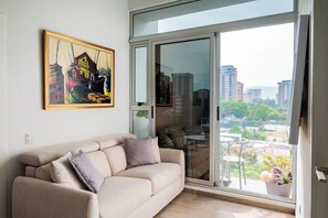 Un salon moderne avec un canapé beige, une porte vitrée donnant sur un balcon et une vue sur la ville.