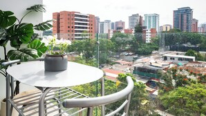 Ein Balkon mit einem Blumentopf und einem Tisch, mit Blick auf eine Stadtlandschaft aus Gebäuden und Grünflächen.