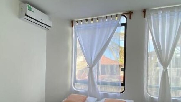 A hotel room with a bed, white curtains, a window, and an air conditioning unit. - Hostal Brisas Del Mar  (Puerto Villamil)
