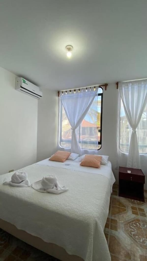 A hotel room with a bed, white curtains, a window, and an air conditioning unit.