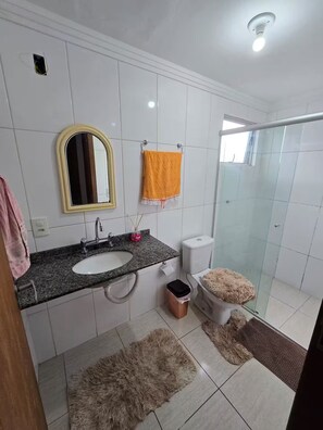 Un baño con lavamanos de encimera de granito, un espejo, un perchero con una toalla anaranjada, un inodoro, un cesto de basura, una zona de ducha y una alfombra mullida en el suelo.