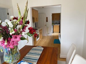 A dining area with a vase of flowers, a striped table runner, and a view into a kitchen with wooden cabinets and a tiled backsplash. - Spacious Family Residence with large south facing gardens close to Eden Project (St Austell)