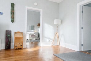 A room with wooden flooring, a standing lamp, a vanity with a mirror, and a door leading to another room. - Village Heritage Suite with Full Kitchen & King Bed (Merrickville)