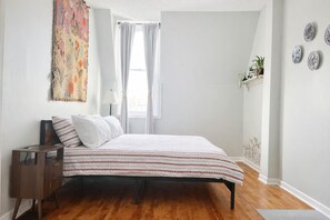 Une chambre à coucher comprenant un lit, une table de chevet, une fenêtre avec des rideaux, une étagère murale ornée de plantes et une assiette décorative accrochée au mur.