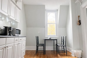 A kitchen with white cabinets, a microwave, a coffee maker, a window, and a table with chairs. - Village Heritage Suite with Full Kitchen & King Bed (Merrickville)