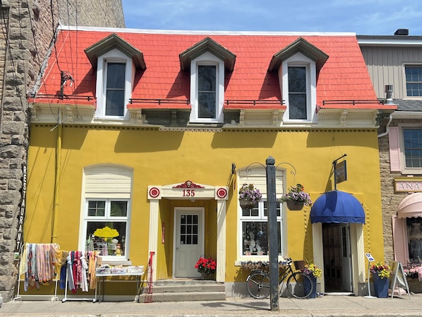 Un bâtiment jaune avec un toit rouge, portant le numéro 135, dont la devanture est ornée de fleurs suspendues et devant lequel est garé un vélo.