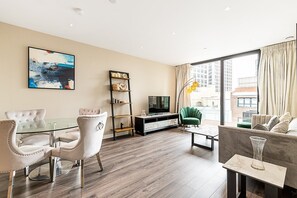 Ein modernes Wohnzimmer mit Glastisch, zwei Polstersesseln, einem grünen Sessel, einem Fernseher, einem Bücherregal und einem großen Fenster mit Blick auf die Stadt.