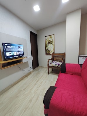 A room with a TV on a wooden shelf, a wooden chair with a floral cushion, a red sofa, and a painting on the wall.