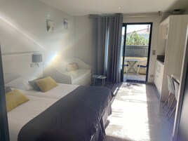 Habitación de hotel con dos camas, una mesita, un sofá, un balcón con mesa y sillas, y una kitchenette.