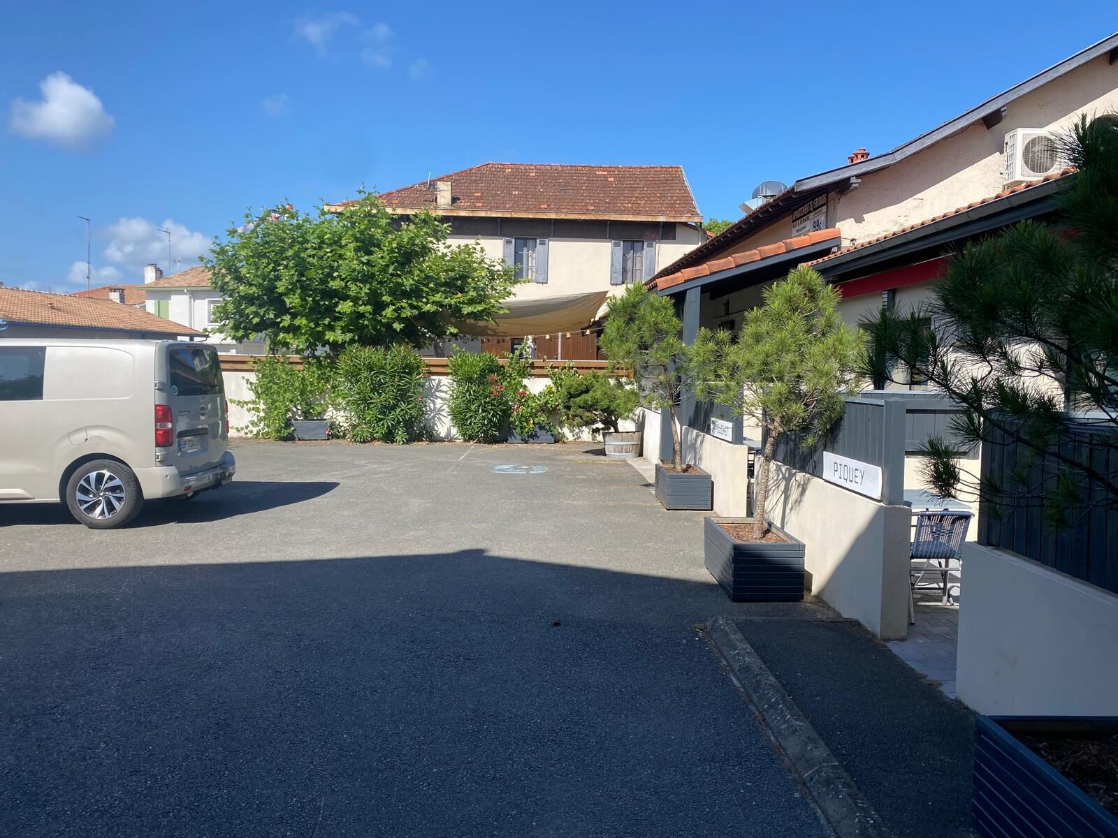 A parking area with a van, a building with a tiled roof, and a sign that reads 'PIQUEY'.