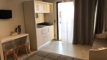 Un espacio habitable compacto con kitchenette, televisor montado en la pared, escritorio con sillas y cama con cabecera.